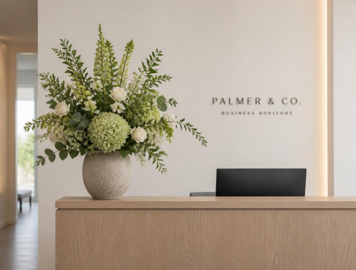 Signature arrangement — a tall 28-inch floral arrangement in a stone vessel on the same light oak reception desk in the same neutral lobby — reference visual shown from the same straight-on angle at desk height.